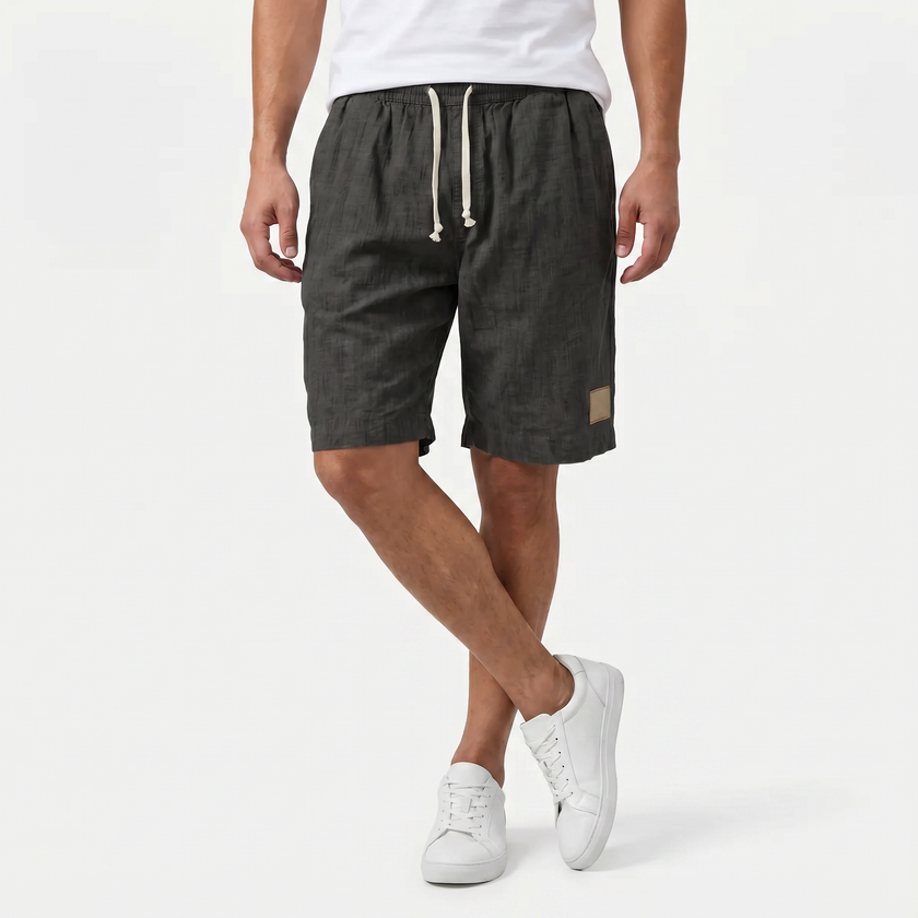 Men's linen shorts by FJØRNA with elastic waist and drawstring, shown from the front with crossed legs on a summer beach style.